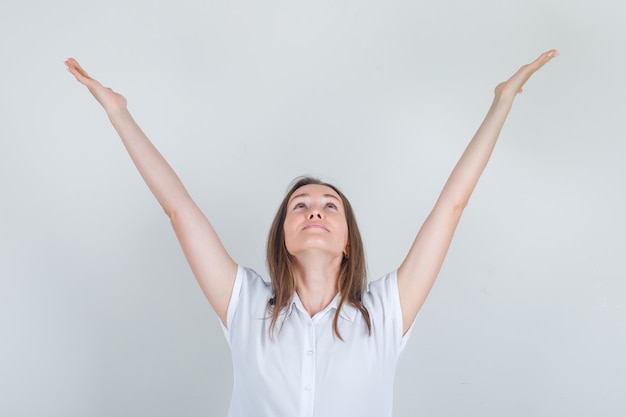 Бесплатное фото Молодая женщина смотрит вверх с поднятыми руками в белой футболке и выглядит веселой