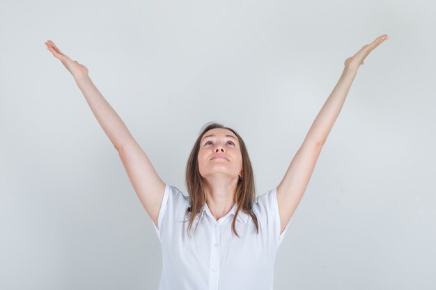 Young woman looking up with raised hands in white t-shirt and looking cheerful