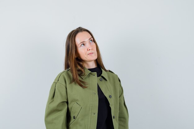 Young woman looking upper left while thinking in green jacket and looking thoughtful , front view.