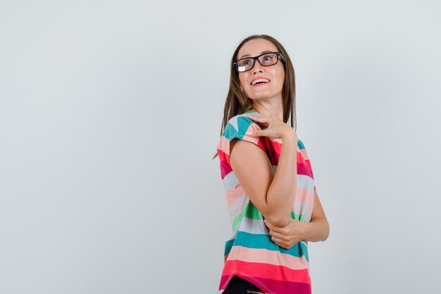 Young woman looking upward in t-shirt, pants, glasses and looking hopeful , front view.