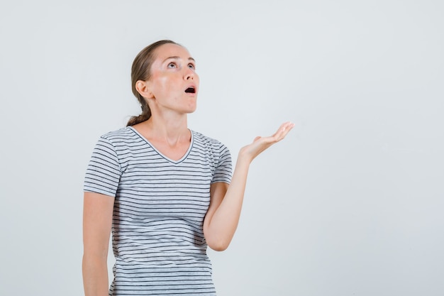 Young woman looking upward with open palm in striped t-shirt and looking astonished. front view.