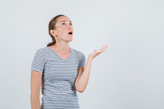 Young woman looking upward with open palm in striped t-shirt and looking astonished. front view.