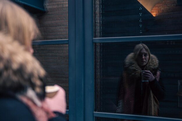 Молодая женщина зимой смотрит на свое отражение в окне