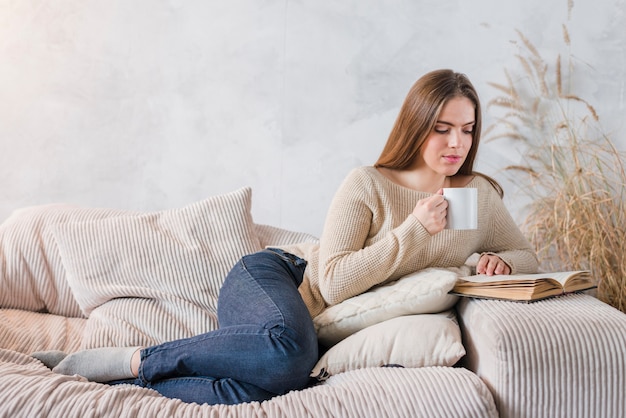 커피 컵을 손에 들고 책을 읽고 침대에 누워있는 젊은 여자