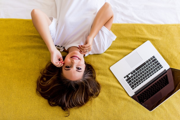 Free photo young woman lying in bed with laptop from above. top view.