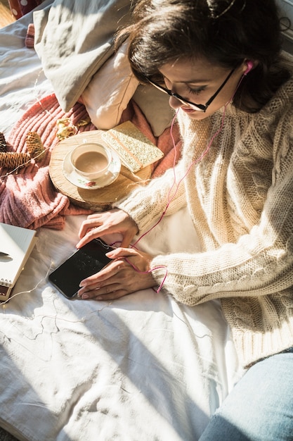 携帯電話とコーヒーでベッドに横たわっている若い女性