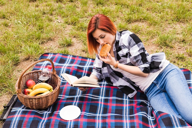 Free photo young woman lying on blanket eating puff pastry reading book