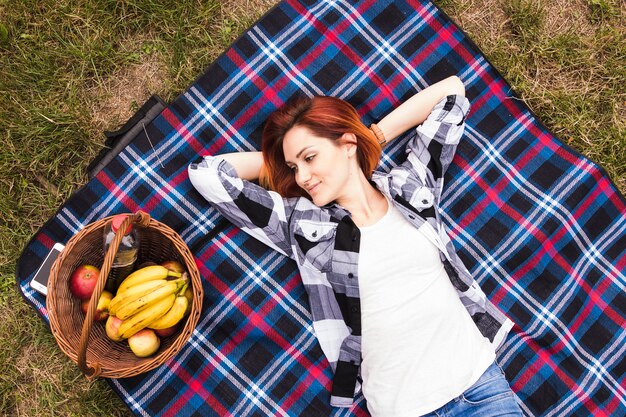 Молодая женщина, лежа на одеяло, глядя на корзину с фруктами и бутылкой
