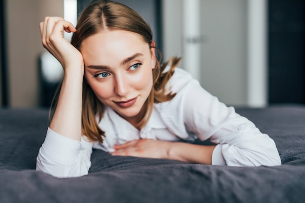 Free photo young woman lying at the end of the bed underneath the quilt and smiling, with her head resting upon her hand with the other in her hair