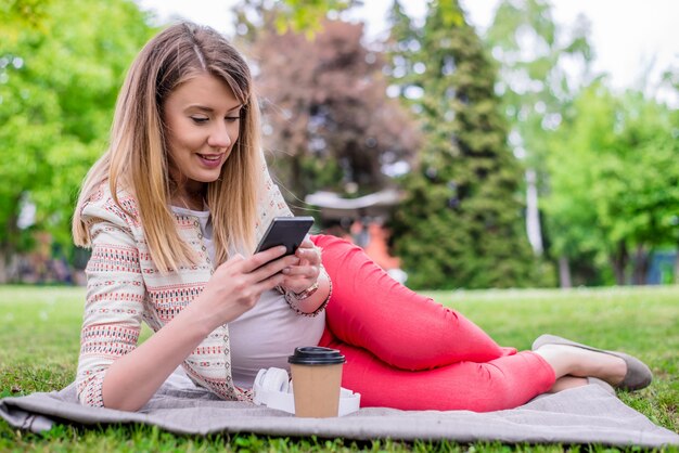 Young woman lying in fresh spring grass listening to music on her mobile phone smiling with pleasure. Young female expecting child having rest outside in public garden. Pregnant woman surfing internet on cell phone.