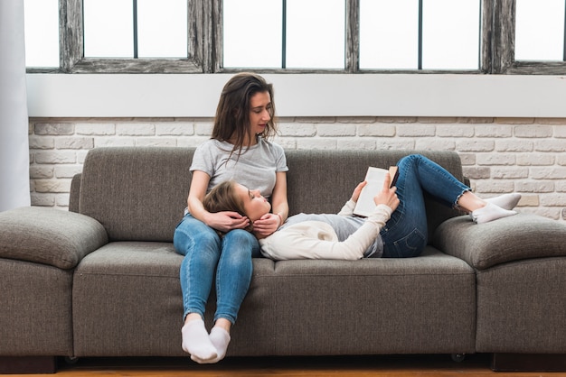 Free photo young woman lying on her girlfriend's lap reading the book on sofa