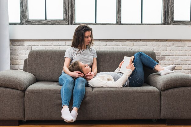 Young woman lying on her girlfriend's lap reading the book on sofa