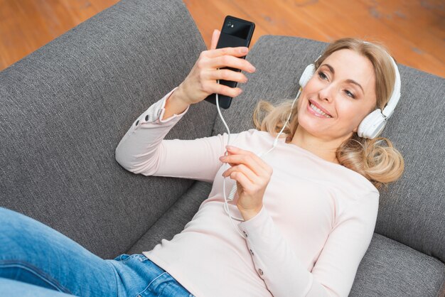 Молодая женщина, лежа на диване, слушать музыку с наушниками с смартфона