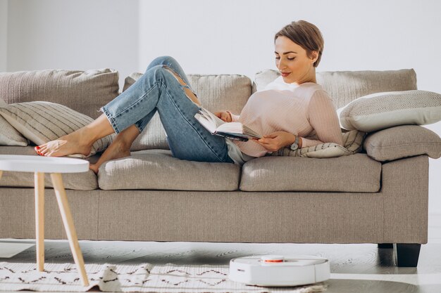 Молодая женщина, лежа на диване и читающая книгу, пока робот-пылесос делает работу по дому