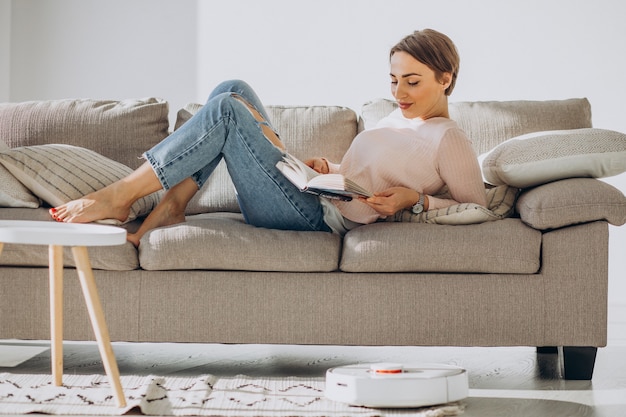 로봇 진공 청소기가 집안일을하는 동안 소파에 누워 책을 읽는 젊은 여자