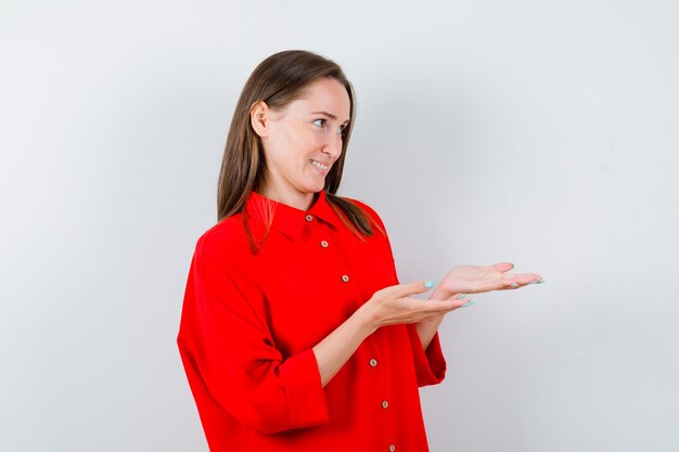 Young woman making giving or receiving gesture in red blouse and looking cheery , front view.