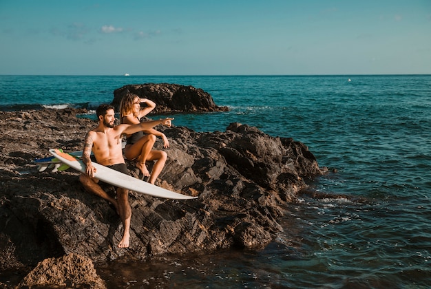 Young woman and man pointing to side with surf boards on rock near sea