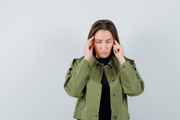 Young woman massaging her temple in green jacket,black shirt and looking stressful , front view.