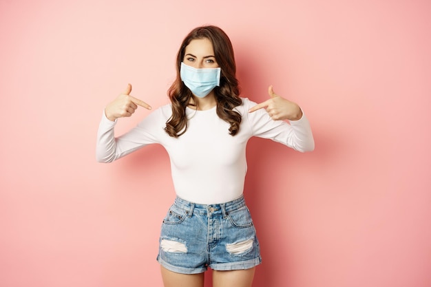 Young woman in medical face mask pointing fingers at herself showing logo or banner smiling at camer...