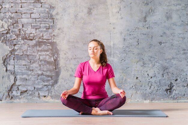 Young woman meditating in lotus pose against damaged grey wall