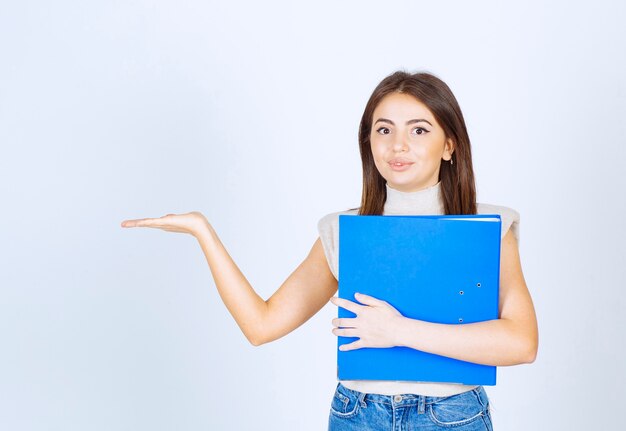 young woman model holding a blue folder and showing a hand over white wall.