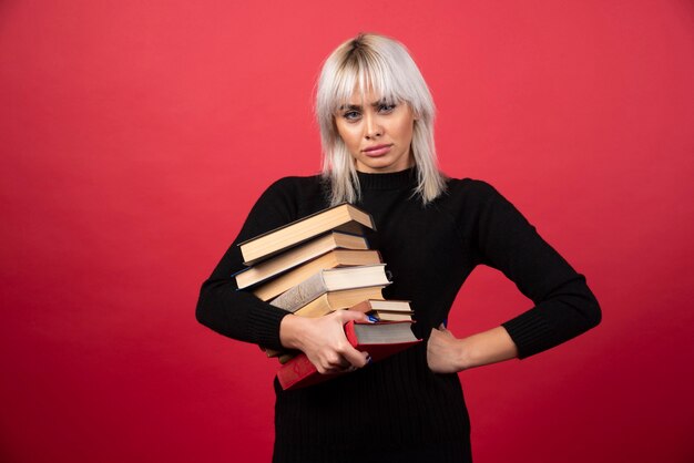 Young woman model holding a lot of books on a red wall. 