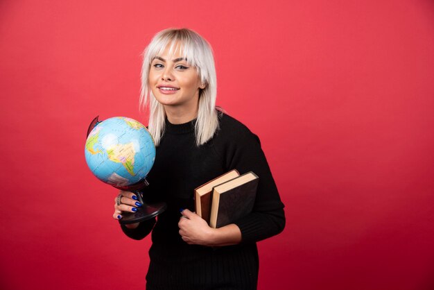 Free photo young woman model posing with a books and a earth globe on a red wall.