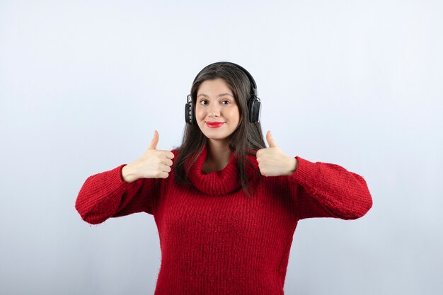 엄지 손가락을 보여주는 헤드폰으로 빨간 스웨터에 젊은 여자 모델