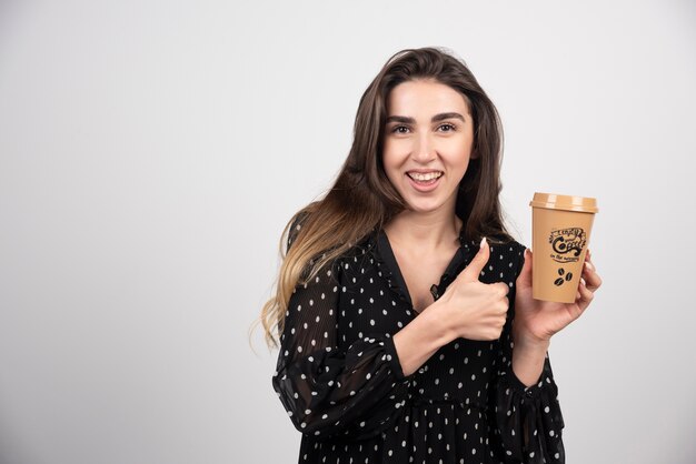 Модель молодой женщины показывает палец вверх и держит чашку кофе