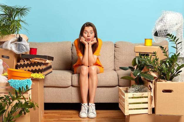 Young woman moves in new bought apartment, poses on couch with shocked expression