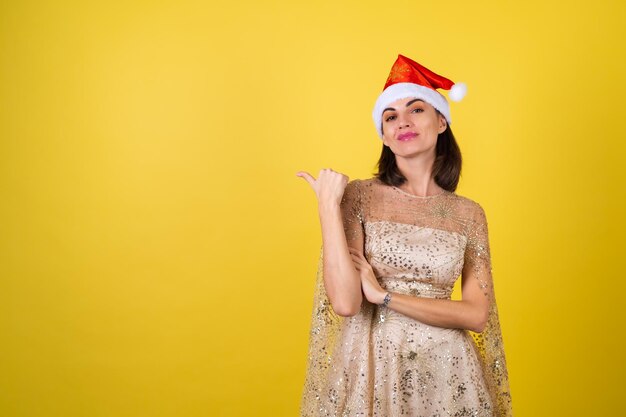 Young woman in new year's eve evening shiny sparkling beige dress and santa hat celebrates the  holidays, points a finger to the left