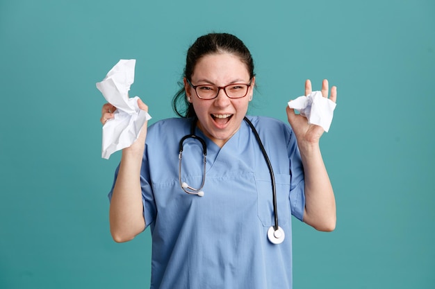 Молодая женщина-медсестра в медицинской форме со стетоскопом на шее комкает бумагу в гневе и кричит с агрессивным выражением лица, стоя на синем фоне