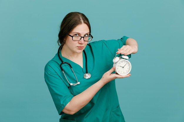 Young woman nurse in medical uniform with stethoscope around neck holding alarm clock looking at camera with serious face standing over blue background