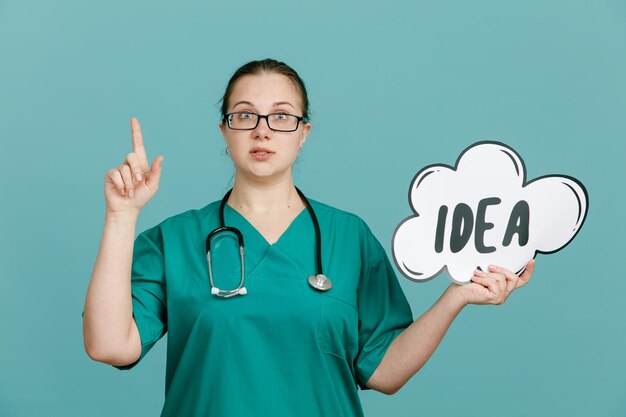 Молодая женщина-медсестра в медицинской форме со стетоскопом на шее, держащая пузырчатую речь с идеей слова, выглядит уверенно, показывая указательный палец, стоящий на синем фоне