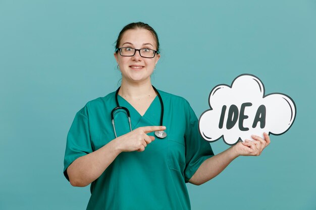 Молодая женщина-медсестра в медицинской форме со стетоскопом на шее держит пузырчатую речь со словесной идеей, указывая указательным пальцем на нее, удивленно улыбаясь, стоя на синем фоне