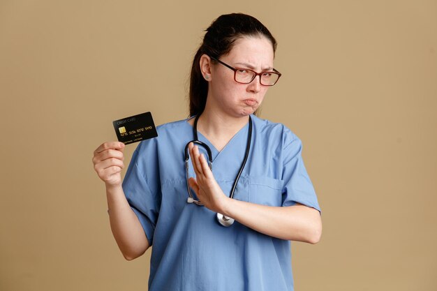 Молодая женщина-медсестра в медицинской форме со стетоскопом на шее, держащая кредитную карту, недовольно хмурится, делая знак "стоп" с открытой рукой, отказываясь стоять на коричневом фоне