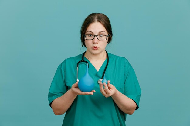 Young woman nurse in medical uniform with stethoscope around neck holding enema looking amazed and surprised standing over blue background