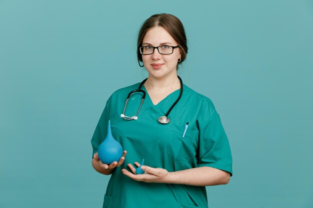 Young woman nurse in medical uniform with stethoscope around neck holding enema looking at camera smiling confident standing over blue background