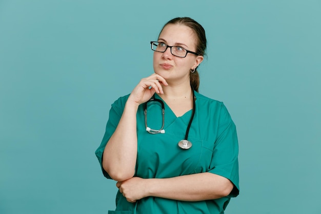파란색 배경 위에 서 있는 턱에 손을 대고 생각에 잠긴 표정으로 옆을 바라보고 목에 청진기를 두른 의료복을 입은 젊은 여성 간호사