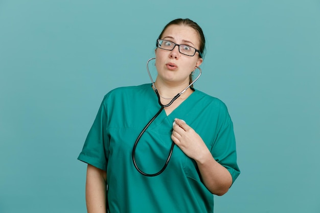 Молодая женщина-медсестра в медицинской форме со стетоскопом на шее смотрит в камеру, пораженная и удивленная стоя на синем фоне
