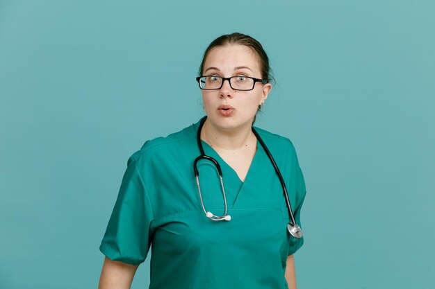 Молодая женщина-медсестра в медицинской форме со стетоскопом на шее смотрит в камеру, растерянно и удивленно стоя на синем фоне
