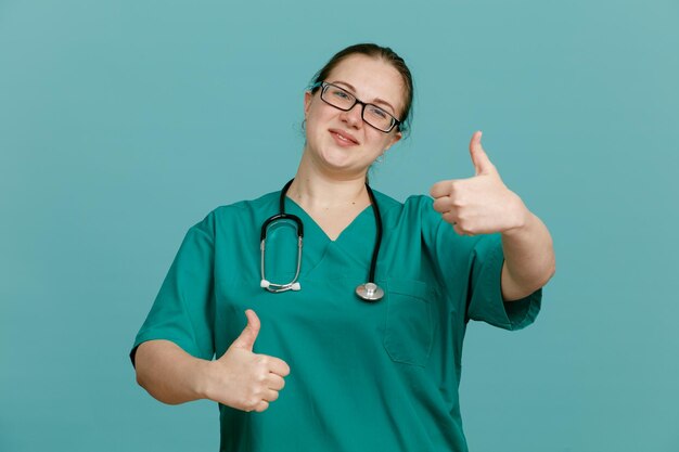 Молодая женщина-медсестра в медицинской форме со стетоскопом на шее смотрит в камеру, счастливая и позитивно улыбаясь, весело показывая большой палец обеими руками, стоя на синем фоне