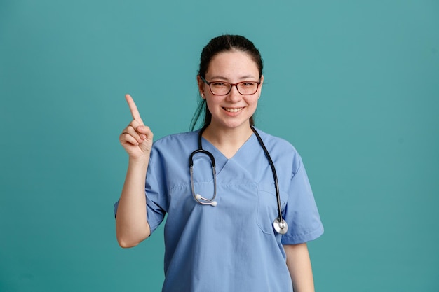 Молодая женщина-медсестра в медицинской форме со стетоскопом на шее смотрит в камеру, счастливая и позитивно улыбаясь, уверенно показывая указательный палец, стоящий на синем фоне