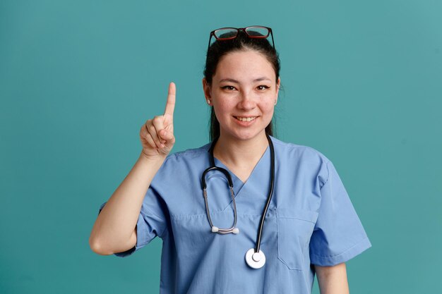 Young woman nurse in medical uniform with stethoscope around neck looking at camera happy and positive smiling showing number one with index finger standing over blue background