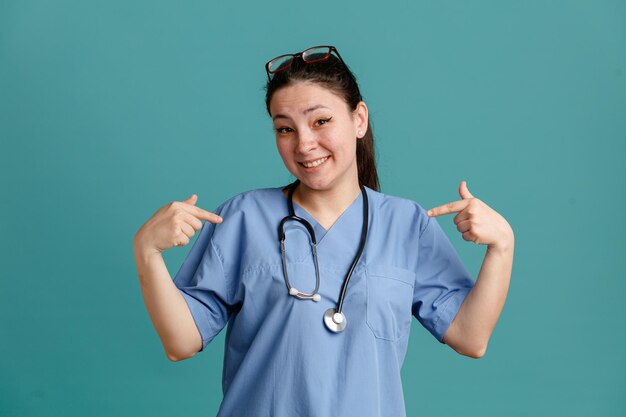 young woman nurse in medical uniform with stethoscope around neck looking at camera happy and selfsatisfied pointing at herself standing over blue background