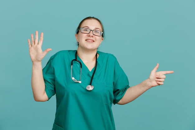 Молодая женщина-медсестра в медицинской форме со стетоскопом на шее смотрит в камеру, весело улыбаясь, показывая номер пять с открытой ладонью, указывающей указательным пальцем в сторону