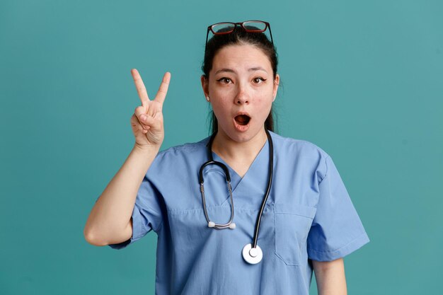 목에 청진기가 달린 의료복을 입은 젊은 여성 간호사가 파란 배경 위에 손가락이 서 있는 2번을 보여주는 카메라를 보고 놀랐습니다.