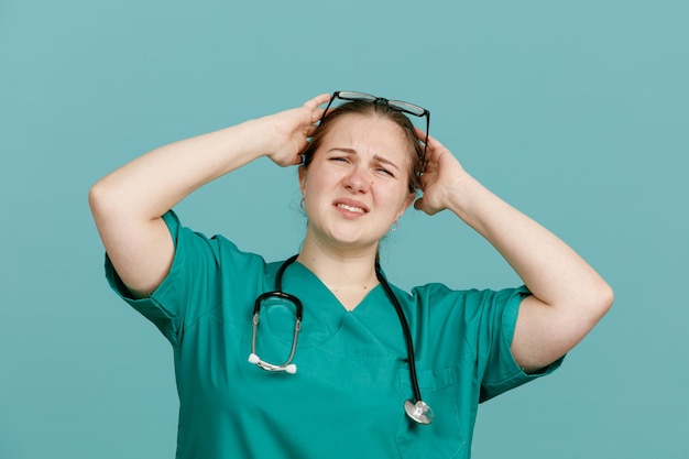 Молодая женщина-медсестра в медицинской форме со стетоскопом на шее выглядит сбитой с толку, держась за голову за ошибку, стоя на синем фоне