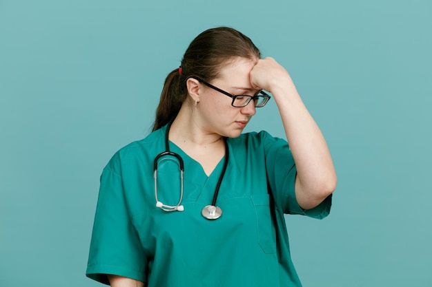 Young woman nurse in medical uniform with stethoscope around neck looking tired and overworked holding hand on her forehead standing over blue background