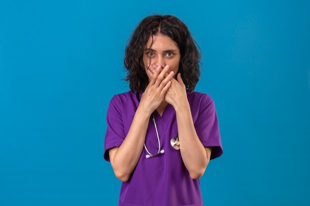 Young woman nurse in medical uniform and with stethoscope looking surprised covering her mouth with hands standing